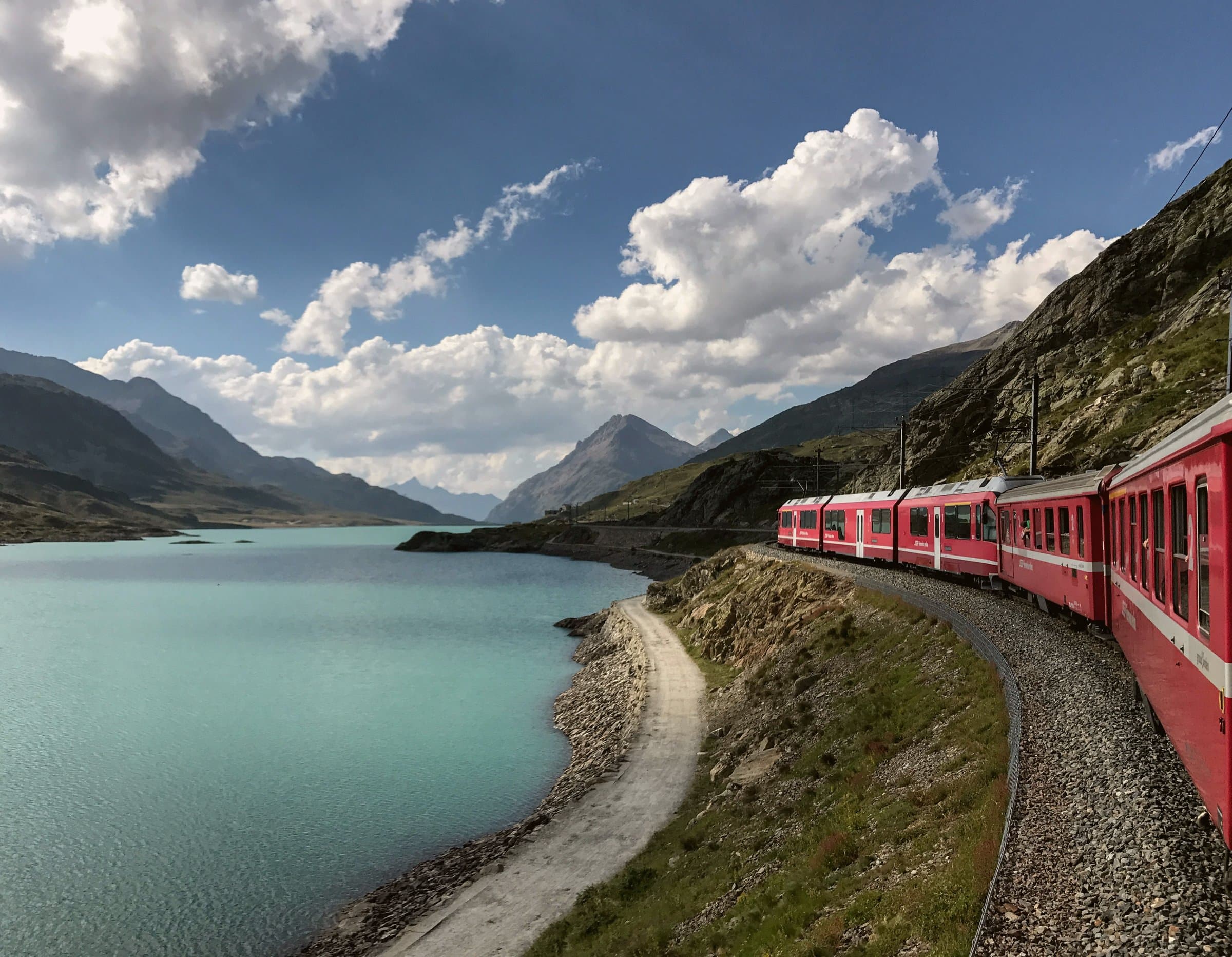 A red Rhaetian Railway train winding beside a turquoise alpine lake in the Swiss Alps