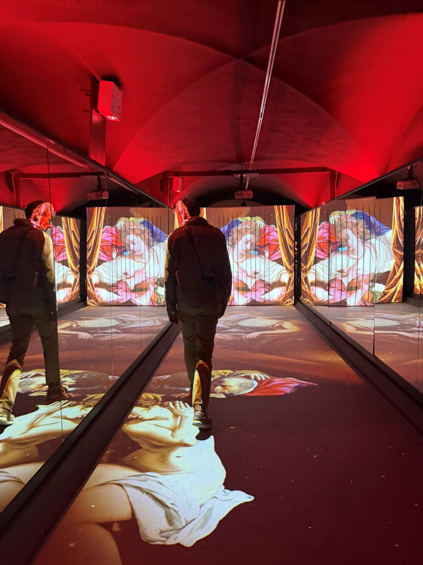 A visitor in a red-lit room where paintings are projected across the walls and floor