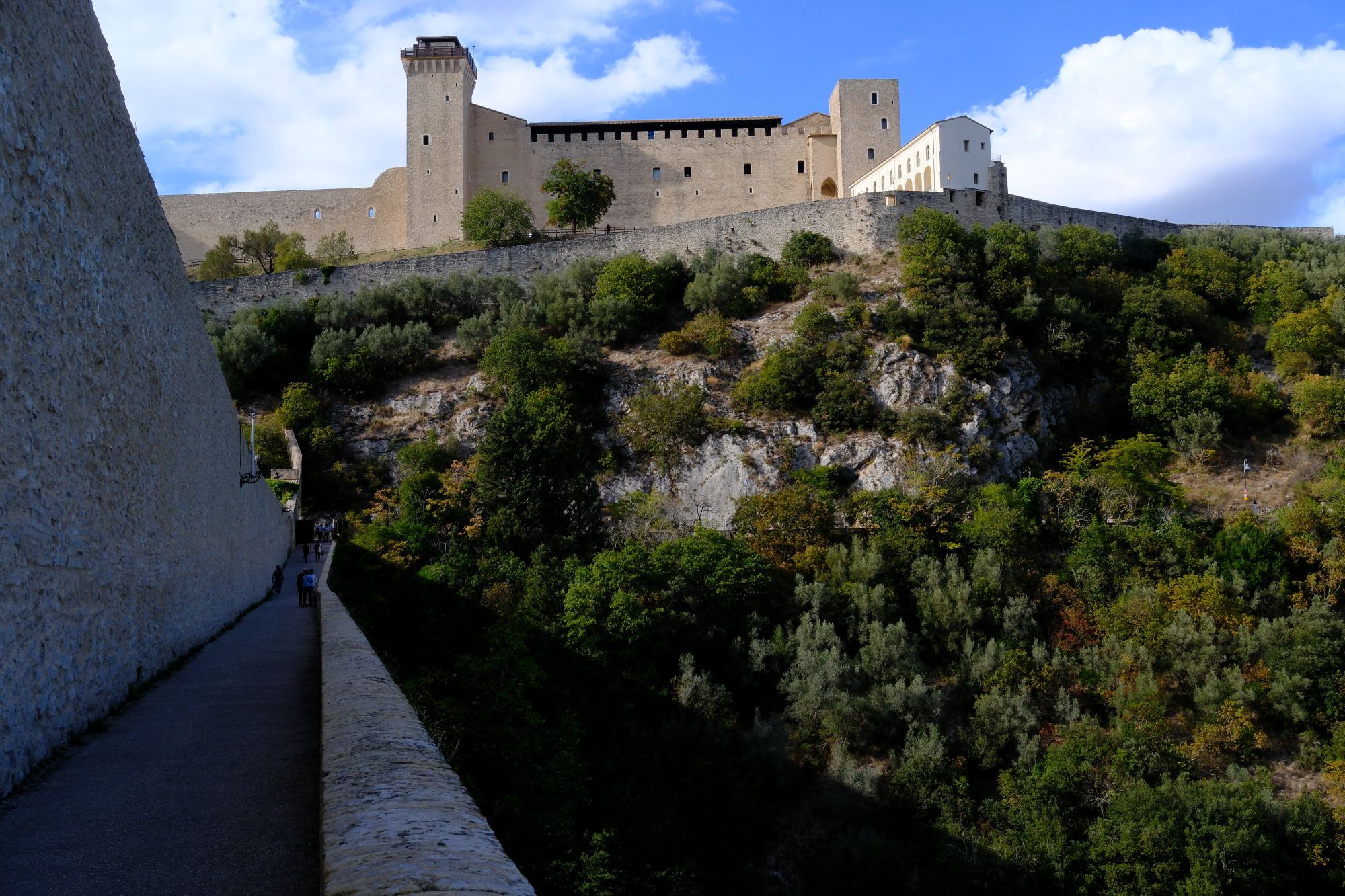The walls of the Rocca Albornoziana above a wooded slope, with a path along the rampart