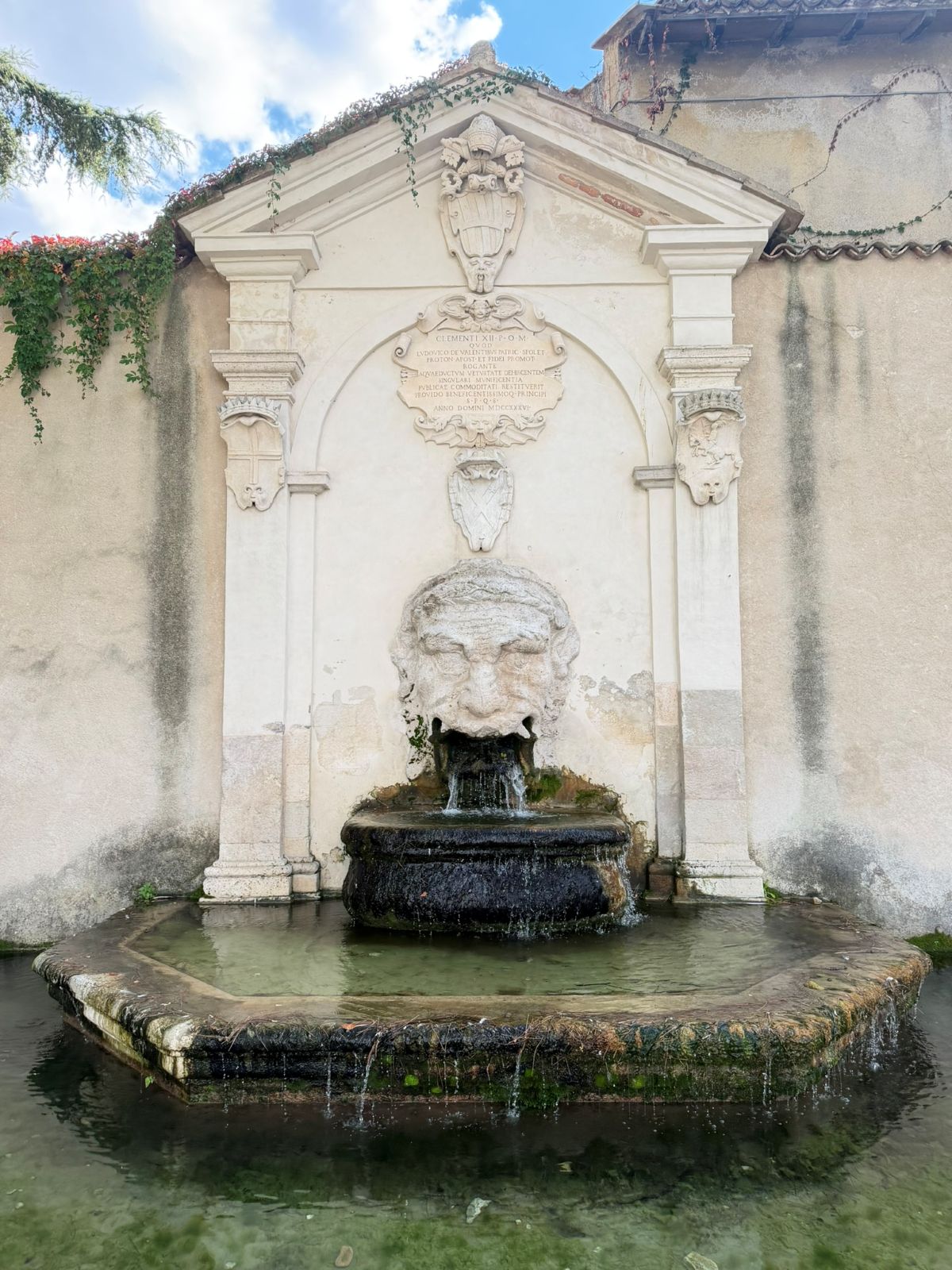 A stone wall fountain in Spoleto, with a carved mask pouring into the basin
