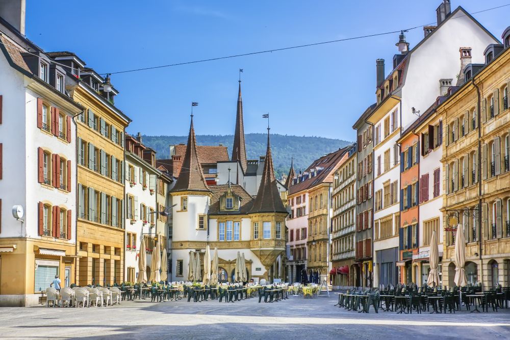 Café tables in a square of yellow stone houses in Neuchâtel, with pointed turrets and a wooded hill behind